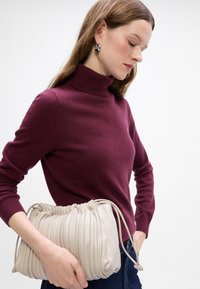 Beige Faltenhandtasche aus weichem Material, mit einem Kordelverschluss und strukturiertem Oberflächen, gehalten neben einem burgunderfarbenen Rollkragenpullover.