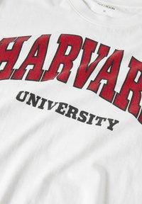 White cotton t-shirt with bold red "HARVARD" and black "UNIVERSITY" text. Crew neckline, soft texture, standard short sleeve design.