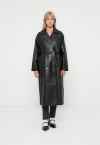 Svart faux lær dobbeltspent trenchcoat med belte, brede reverser og lange ermer; glatt tekstur og knelengde.