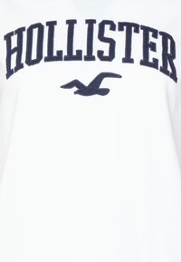 White t-shirt featuring navy blue embroidered "HOLLISTER" text and a seagull logo underneath. Cotton material, standard crew neck design.