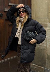 Juoda pūkinė striukė su kapišonu, siūta tekstūra, derinama su smėlio spalvos šaliku ir su leopardo rašto sijonu. Laiko juodą siūtą rankinę.