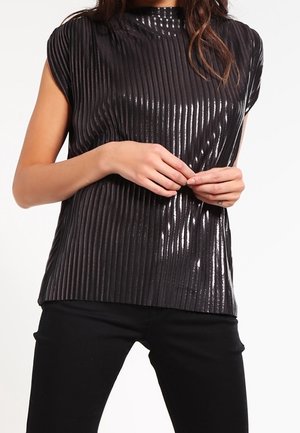 Femme portant un haut noir brillant plissé à manches courtes avec un pantalon noir, les mains délicatement jointes au niveau de la taille.