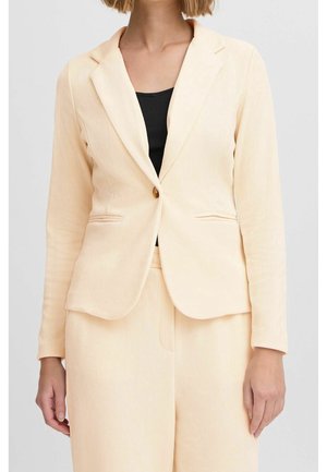 Kvinde iført beige fløjlsblazer og matchende bukser over en sort top, stående mod en ensfarvet lysegrå baggrund.
