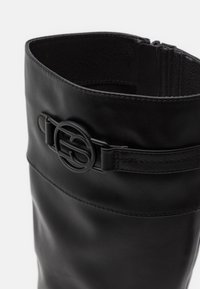 Bota de cuero negro hasta la rodilla con una textura suave, que cuenta con un cierre lateral y un acento de logotipo circular en la correa.