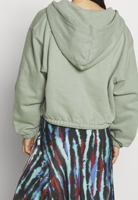 Sudadera de gran tamaño de color verde claro con capucha, dobladillo acanalado y puños elásticos, combinada con una falda de patrón multicolor con franjas rojas, azules y negras.