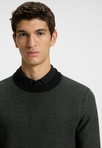 Dark green knit sweater with a textured finish and a black ribbed collar, featuring a round shape and buttoned collar detail.