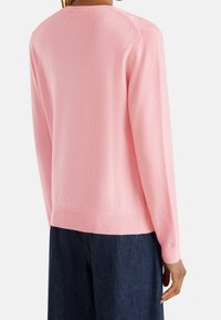 Person klädd i en ljusrosa långärmad tröja och mörkblå jeans, sedd från baksidan mot en enkel vit bakgrund.