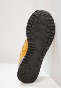 Svart grov gummisula på en gul sneaker vinklad mot vita väggar, som visar djupa spår och en strukturerad greppmönster.