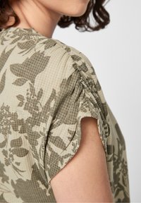 Koszula w kwiatowy wzór w oliwkowym zielonym i beżowym kolorze, wykonana z miękkiej, teksturowanej tkaniny, o luźnym kroju i podwiniętych rękawach.