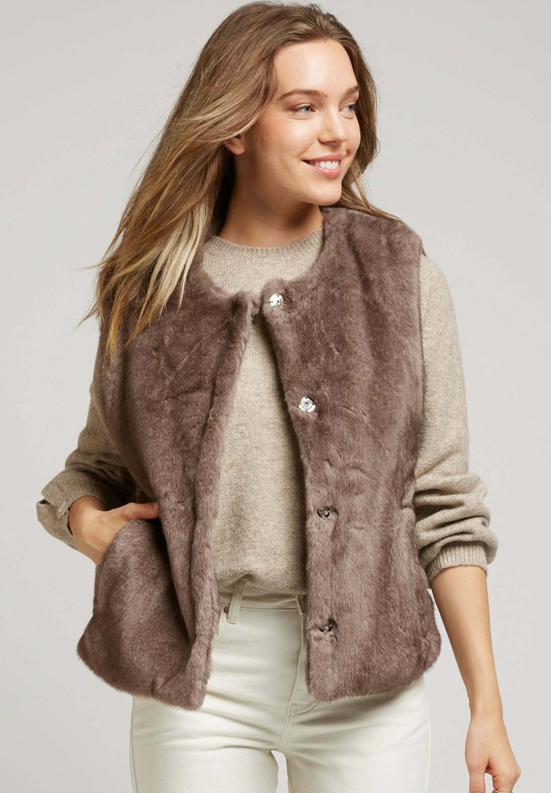 Femme portant un pull beige clair et un pantalon blanc, souriant, avec la main dans la poche d'un gilet en fausse fourrure marron boutonné.