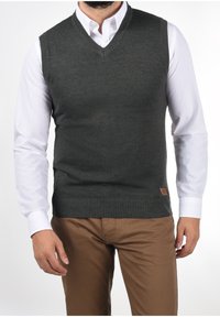 Chaleco de punto gris con escote en V, bordes acanalados y una pequeña etiqueta de cuero marrón, superpuesto a una camisa blanca con cuello y pantalones marrones.