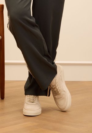 Beige sneakers with textured fabric, flat sole, and lace closure. Worn with dark pants featuring a side stripe. Wooden chair partially visible.