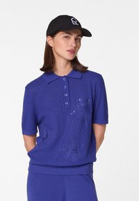 Blaues Poloshirt mit kurzen Ärmeln, das mit gestickten Blumenmustern und Knöpfen verziert ist. Kombiniert mit einer schwarzen Mütze. Weicher Stoff und lässiger Schnitt.