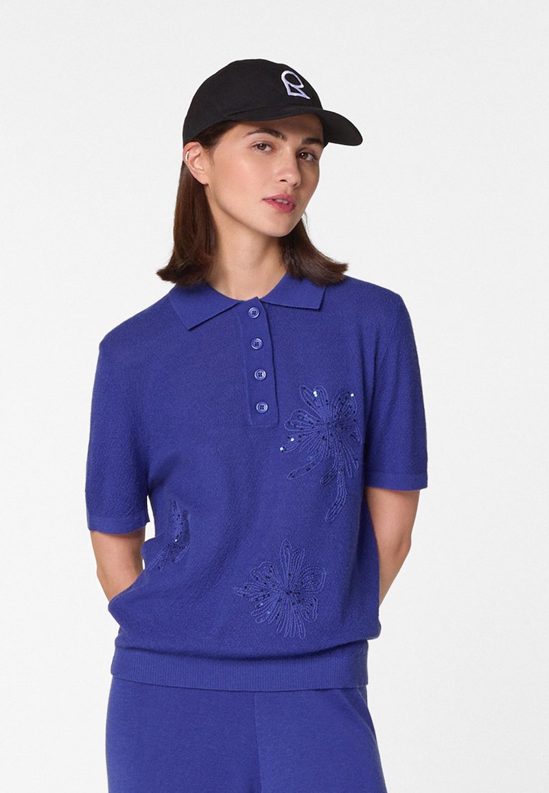 Blaues Poloshirt mit kurzen Ärmeln, das mit gestickten Blumenmustern und Knöpfen verziert ist. Kombiniert mit einer schwarzen Mütze. Weicher Stoff und lässiger Schnitt.