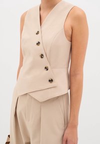 Beige bezramenný top s výstřihem do V, s asymetrickým lemem a pěti knoflíky z želvovinového vzoru vpředu, vyrobený z hladkého materiálu.