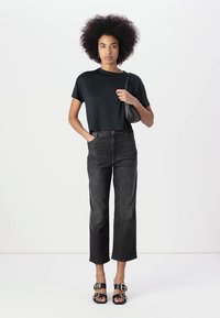 Μαύρο κοντό μπλουζάκι, ψηλόμεσα μαύρα τζιν με ίσια γραμμή και μαύρες σαγιονάρες με αγκράφες. Μοντέλο που κρατά μια μικρή μαύρη τσάντα.