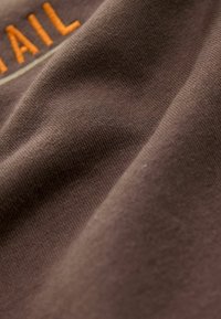 Gros plan sur un tissu marron avec une texture tricotée visible et une partie du texte brodé orange "TAIL" dans le coin supérieur gauche.