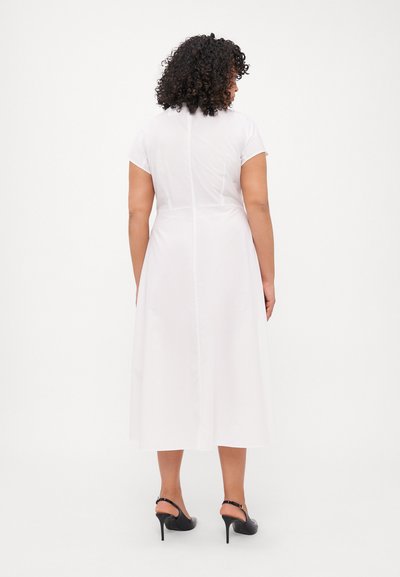 Robe blanche mi-longue avec des manches courtes, texture lisse, et une couture au milieu du dos. Portée avec des sandales à talons noires.