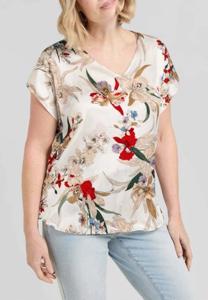 Donna che indossa una blusa bianca a maniche corte con fiori rossi, beige e viola, abbinata a jeans azzurri su uno sfondo neutro.