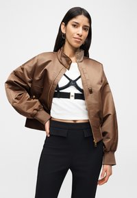 Γυναίκα με μακριά σκούρα μαλλιά φοράει καφέ σατέν bomber μπουφάν πάνω από λευκό κοντό τοπ και μαύρο harness, συνδυασμένο με μαύρο παντελόνι ψηλόμεσο.