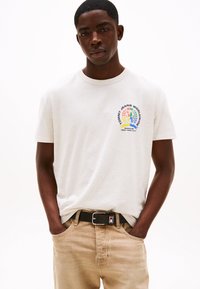 Lys grå bomull t-skjorte med en fargerik grafisk logo på venstre bryst. Matchet med beige jeans og et svart belte med metallspenne.