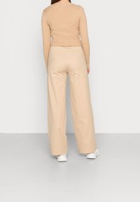 Femme portant un pantalon large beige, un haut à manches longues beige à côtes, et des baskets blanches, se tenant face à un mur blanc.