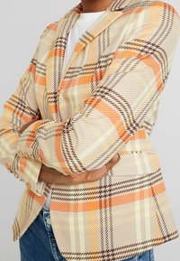 Osoba w pomarańczowej, beżowej i brązowej marynarce w kratę, nosząca białą bluzkę, z założonymi rękami, w połączeniu z niebieskimi dżinsami.