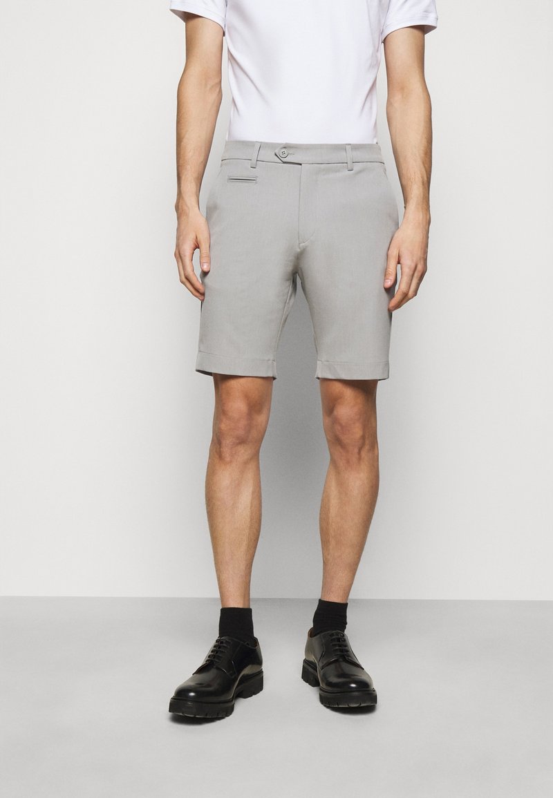 Homme portant un short gris clair sur mesure, des chaussettes noires, des chaussures habillées noires et une chemise blanche à manches courtes, debout devant un fond uni.