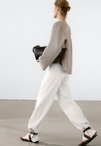 Lichtgrijze linnen trui, losse witte broek en bruine sandalen. Houdt een gestructureerde zwarte clutch vast. Eenvoudige, moderne silhouet.