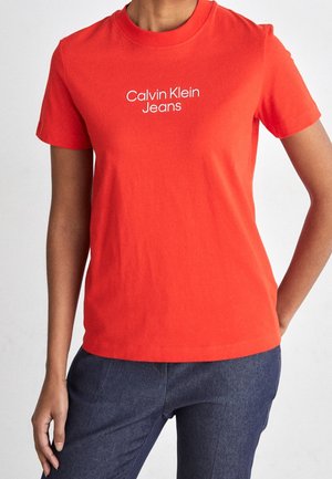 Osoba nosząca jasnoczerwoną koszulkę z krótkim rękawem z tekstem "Calvin Klein Jeans" i ciemnoniebieskie dżinsy na jednolitym tle.