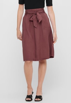 Vrouw die een knie-lange bruine rok met taillestrik draagt, gecombineerd met een zwart topje met korte mouwen en zwarte sandalen met open teen, staand tegen een effen achtergrond.