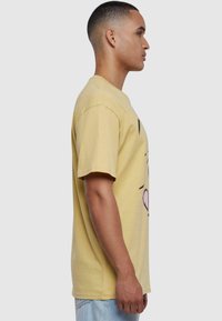 Gul bomull T-skjorte med stort grafisk design på forsiden, kort erm, og klassisk rund hals, vist på en modell fra siden.