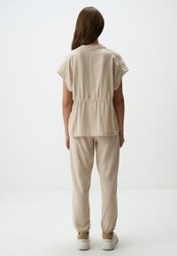 Beige, kurzärmliges Oberteil mit geraffter Taille, kombiniert mit locker sitzenden beigen Hosen. Das Outfit zeichnet sich durch ein minimalistisches Design und eine glatte Textur aus.