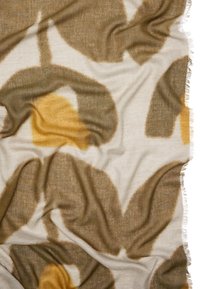 Koc z mieszanki wełny z abstrakcyjnymi wzorami liści w ziemistych odcieniach brązu i musztardy na jasnym, neutralnym tle, z postrzępionymi brzegami.