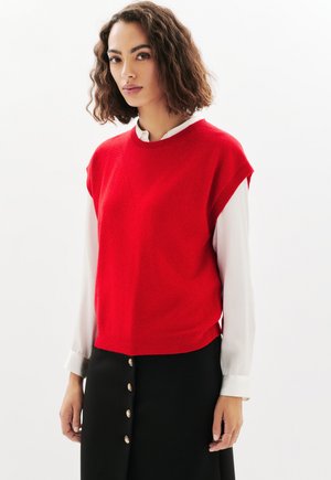 Jonge vrouw met golvend bruin haar die een rode mouwloze trui over een witte lange mouwen shirt en een zwarte knopenrok draagt.