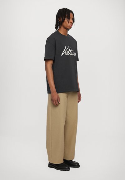Schwarzes Baumwoll-T-Shirt mit einem weißen Schriftzug-Grafik, kombiniert mit beigefarbenen weit geschnittenen Hosen und schwarzen Schuhen mit dicker Sohle.