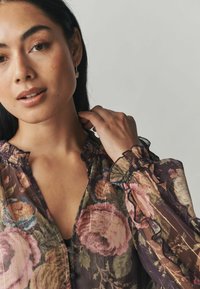 Permatomas gėlių marškinėlis su pūstomis apykakle ir manžetais, turintis tamsų foną, rožinės ir žalios gėlės raštą, bei sagų užsegimą.