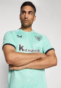 Mann mit verschränkten Armen, der ein hellblaues Fußballtrikot des Athletic Club Bilbao mit schwarzen Akzenten und dem Sponsorlogo von Kutxabank trägt.