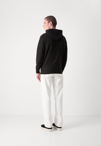 Hanorac negru cu glugă, cu o textură specifică, mâneci lungi și mansete ribbelt, asortat cu pantaloni albi drepti și pantofi negri fără sireturi.