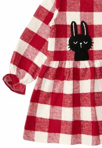 Punainen ja valkoinen ruudullinen mekko, jossa on musta pupu-kuvio. Pitkät hihat, joissa on cuff-yksityiskohtia, ja kerätty vyötärölinja. Pehmeä kangasperhe.