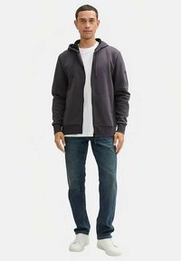 Grå lynlåstrøje med snore, forlommer, kombineret med en hvid T-shirt og blå jeans. Model iført hvide sneakers.