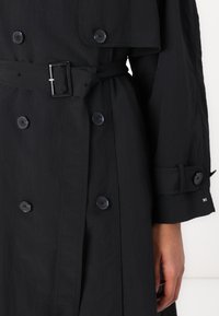 Cappotto nero di trench con design doppiopetto, grandi bottoni, vita con cintura e materiale testurizzato con polsini arrotolati.