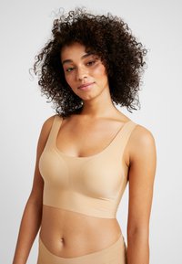Mujer con cabello rizado vistiendo un sujetador beige sin costuras y unas braguitas altas a juego, sobre un fondo liso claro.