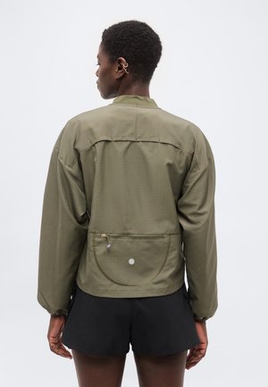 Tagantvaade isikule, kes kannab oliivrohelist jope lukuga tasku ja musti lühikesi pükse, seisab tühja valge tausta ees.