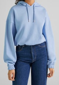 Sudadera con capucha azul claro, de manga larga y puños acanalados, combinada con vaqueros de mezclilla oscuros de cintura alta. Textura suave, corte relajado.