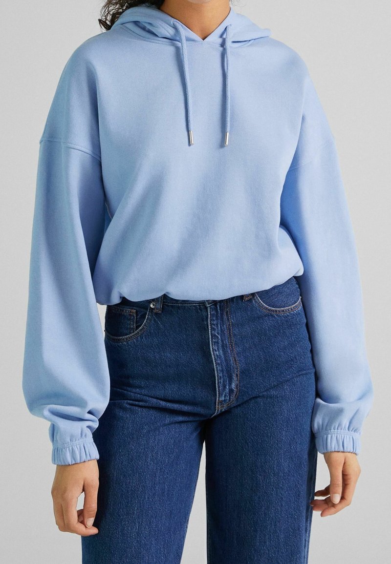 Felpa azzurro chiaro con cappuccio, maniche lunghe e polsini a costine, abbinata a jeans in denim scuro a vita alta. Tessuto liscio, vestibilità comoda.