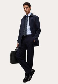 Mężczyzna w granatowym płaszczu, jasnoniebieskiej koszuli, w paskowaną krawat, w spodniach w prążki, czarnych butach, trzymający czarną teczkę, stojący z jedną nogą skrzyżowaną.