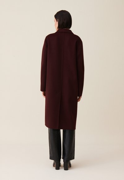 Donna di spalle che indossa un lungo cappotto bordeaux scuro, pantaloni neri a gamba larga e stivali neri con tacco su uno sfondo beige chiaro.