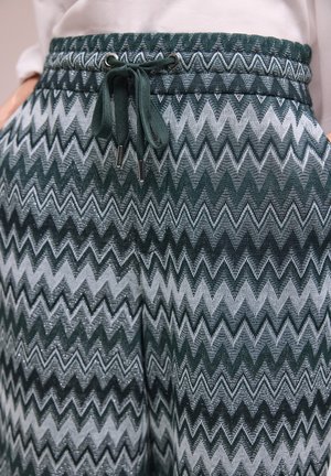 Nahaufnahme von dunkelgrünen und grauen Hosen mit Zickzackmuster, Kordelzugbund und Taschen, getragen zu einem blassen Oberteil.