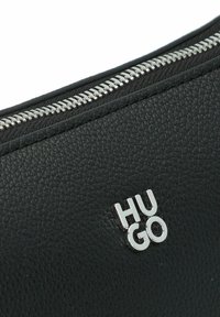 Bolso de cuero negro con acabado texturizado, que presenta un zipper plateado y un logotipo plateado "HUGO" en el lateral.
